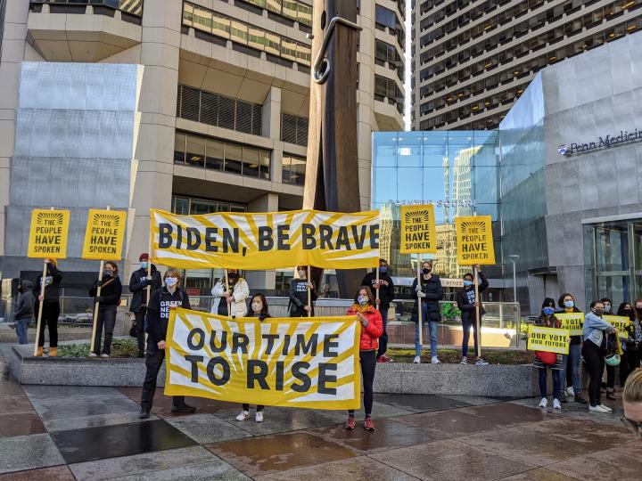 Sunrise Movement protesters stand in front of the clothespin across from City Hall. Their banners read 'Biden, Be Brave', 'Our Time To Rise', while others carry signs reading 'the people have spoken' with a sunrise graphic in the background.