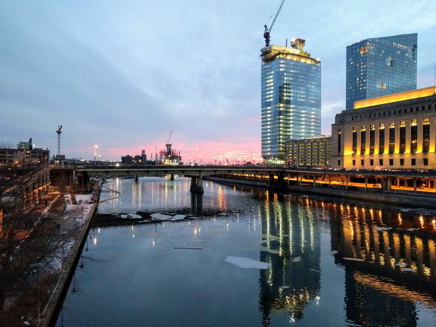 The Schuylkill River in winter. There's snow on the ground, the sun is setting, and the city lights are reflecting in the icy water. The river trail is visible through the trees on the left.