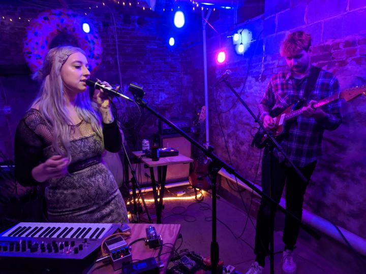 Two musicians performing in an artistically-lit basement. A woman sings into a microphone and a man plays electric guitar.