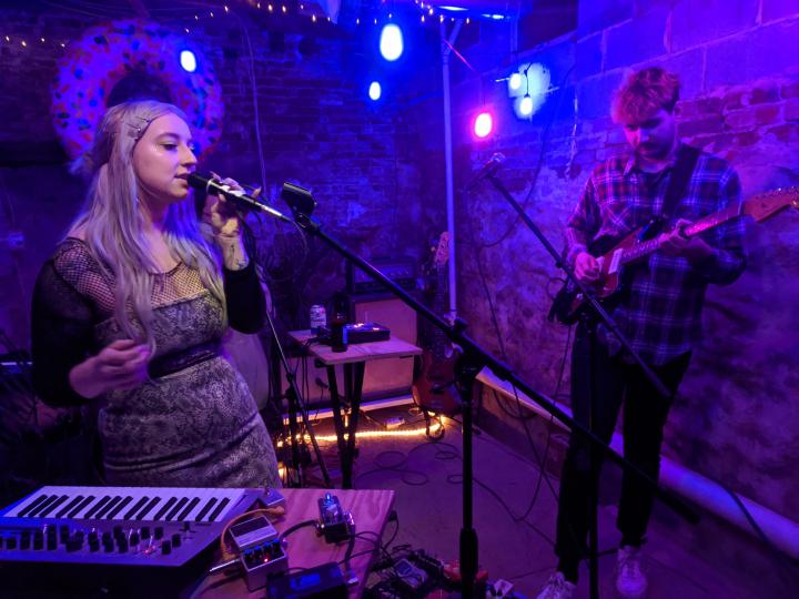 Two musicians performing in an artistically-lit basement. A woman sings into a microphone and a man plays electric guitar.